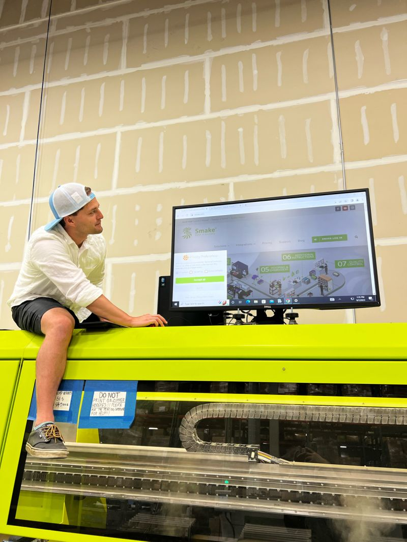 Reif on a factory floor, sitting on a lime-green industrial machine reviewing production software on a monitor